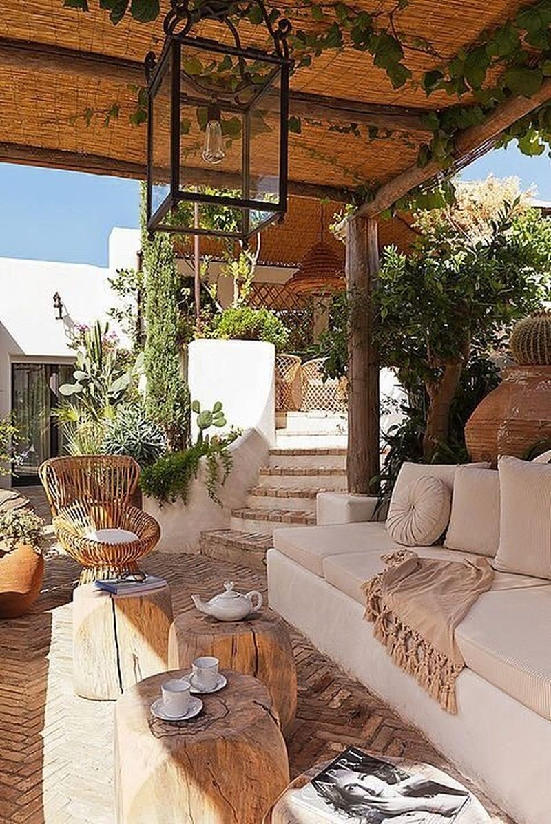 An awesome boho exterior with a hanging lamp, vines on the ceiling, beams table, a rattan chair, light grey sofa to decorate your home exterior design