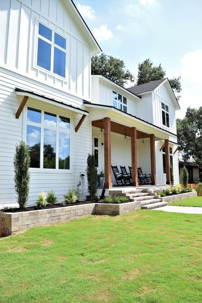 Stunning creekside designs modern farmhouse with white exterior, dark shingle roof, natural wood beams & awning anchors, covered porch with wood ceiling