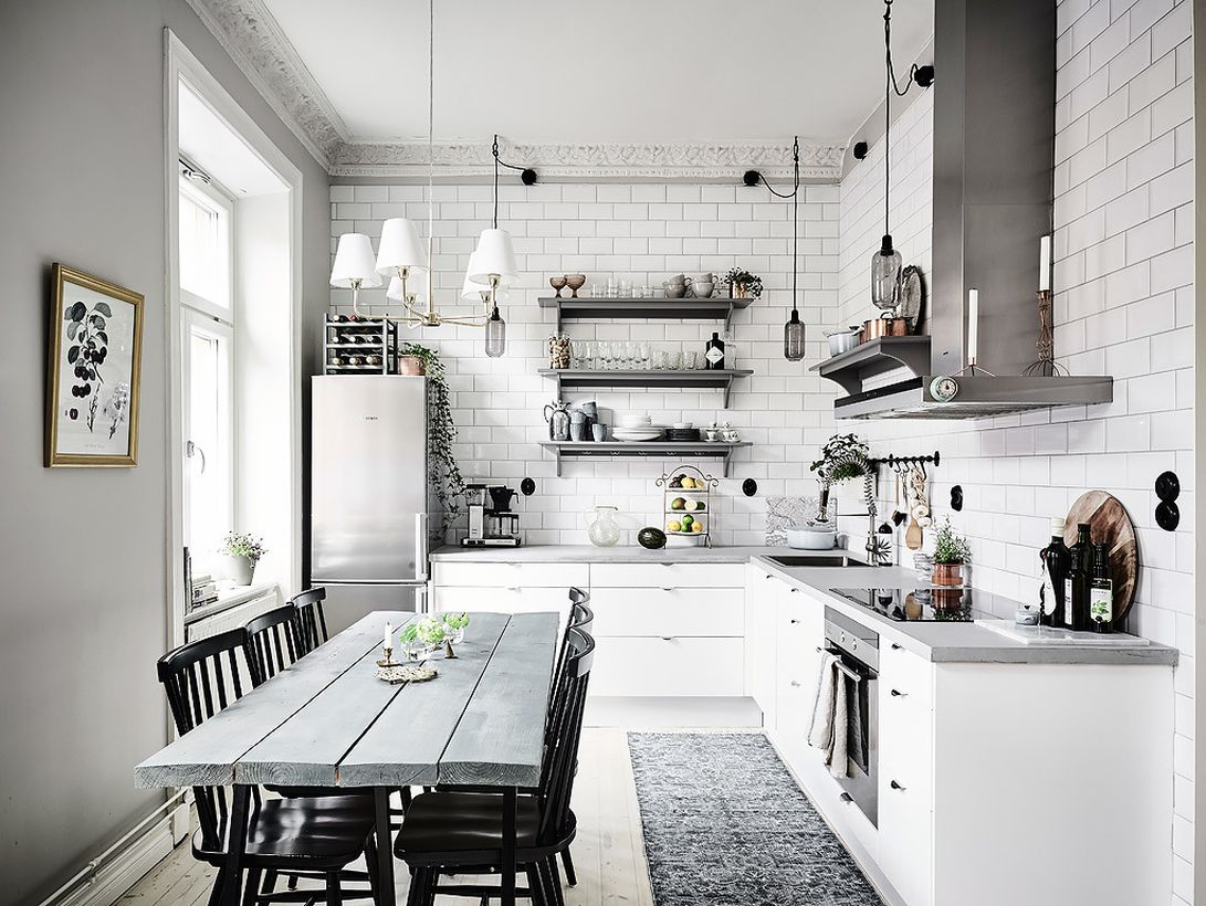 A gorgeous white and grey kitchen design with white brick walls, grey hanging racks to put some kitchen equipment, a grey modern stove, a grey chimney hood cooker, and dining room design