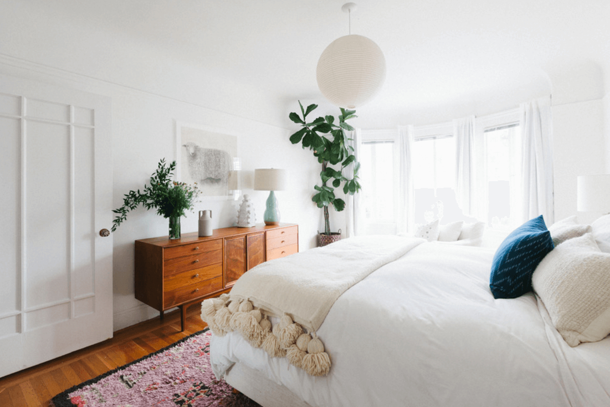 A modern bedroom design with a white sliding door, a wooden bedroom storage, white table lamps, white curtains, and house plants to create a cozy