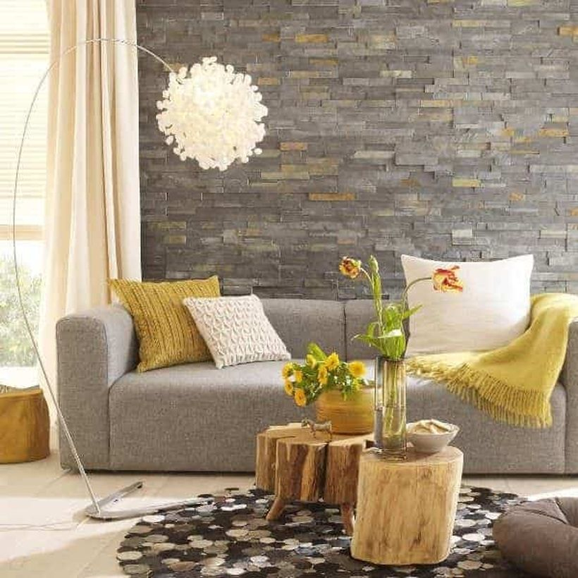 A modern living room design with natural stone brick walls, a window, a beige curtain, a long grey sofa, coffee tables that made of wooden beam and a unique floor lamp to crete to beautify, and house plants