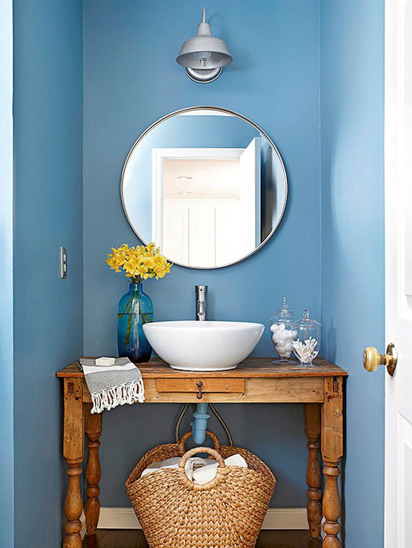 A simple small bathroom decor ideas with blue walls, rustic wooden table for sink, a round mirror, rattan basket to put towels and decorative lighting