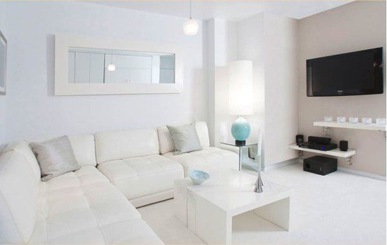 A simple white living room design with white l shaped sofas, a white coffee table, white hanging racks, a table lamp at the corner, and a tv hang on the walls