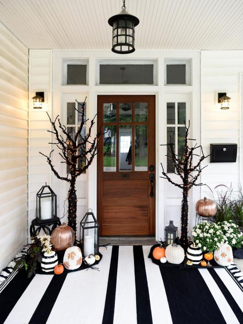 An awesome front porch fall with pumpkins, candle lights, white flower plants, a hanging lamp, lights on the walls, and dried tree lights