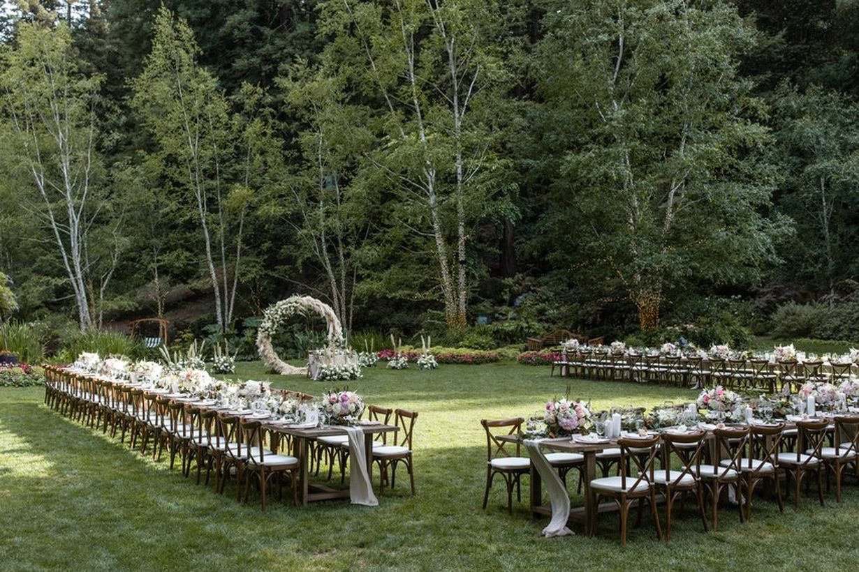 A fabulous outdoor table set for wedding with long wooden tables which is positioned u-shaped, wooden chairs classic, decoration on the table,green grass and background the forest.