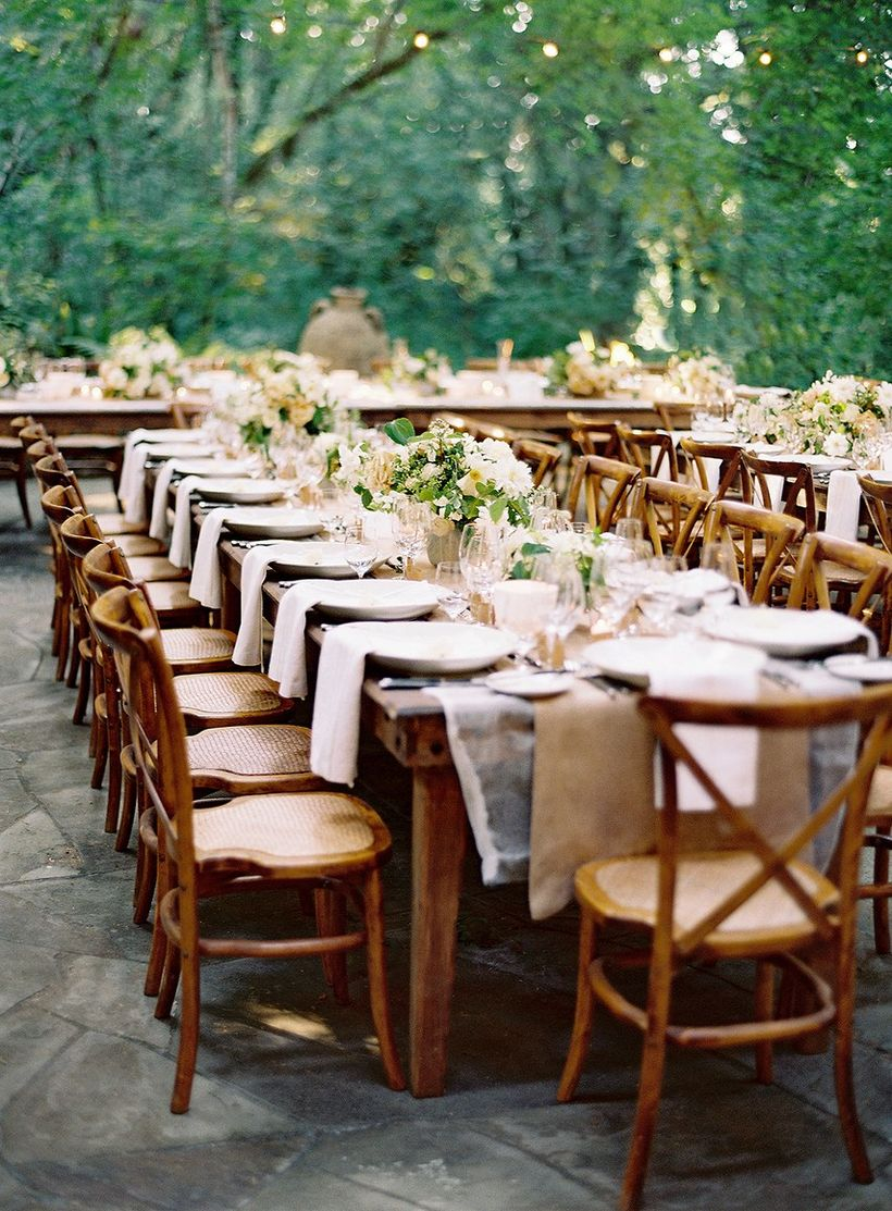 A fabulous outdoor table set for wedding with short wooden table, wooden chairs, white tablecloth and flower decoration on the table.