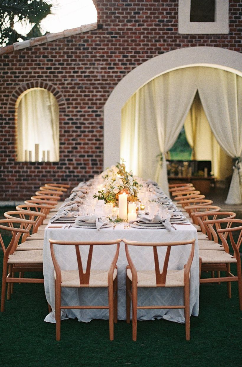 A gorgeous outdoor table set for wedding with long table, white tablecloth, wooden chairs and decoration on the table green grass.