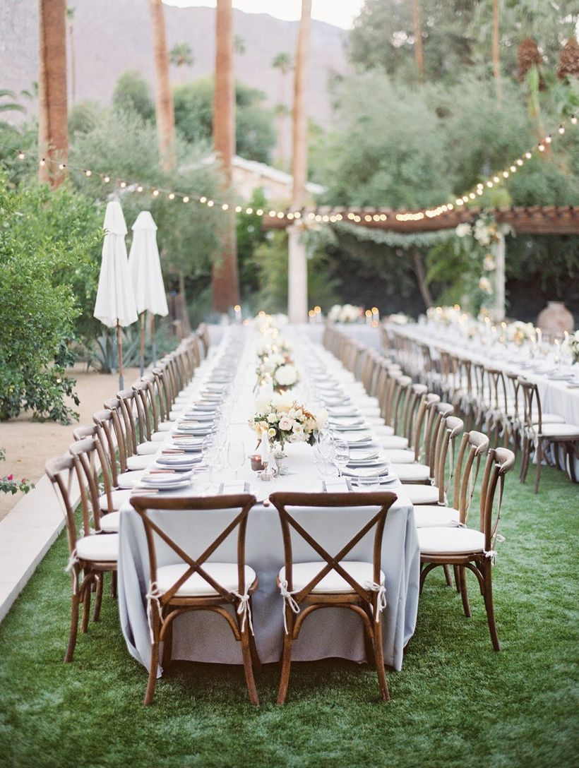 A magnificent outdoor table set for wedding with a long romantic table with floral centerpieces, crisp white dinnerware, green grass and rustic cross-back chairs.