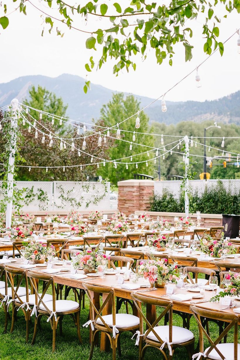 A stunning outdoor table set for wedding with long wooden tables, white tablecloths, wooden chairs, chandeliers and flower decoration on the table.