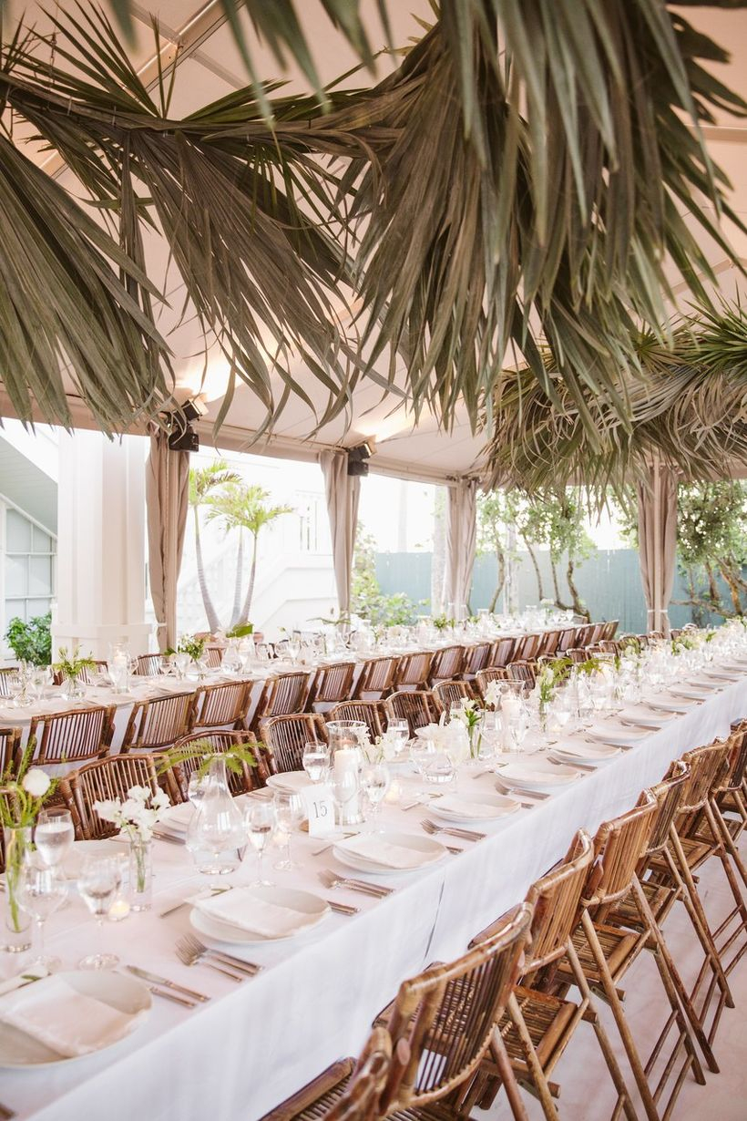 An adorable indoor table set for wedding with long wooden tables, rattan chairs, dried palm leaves and white tablecloth