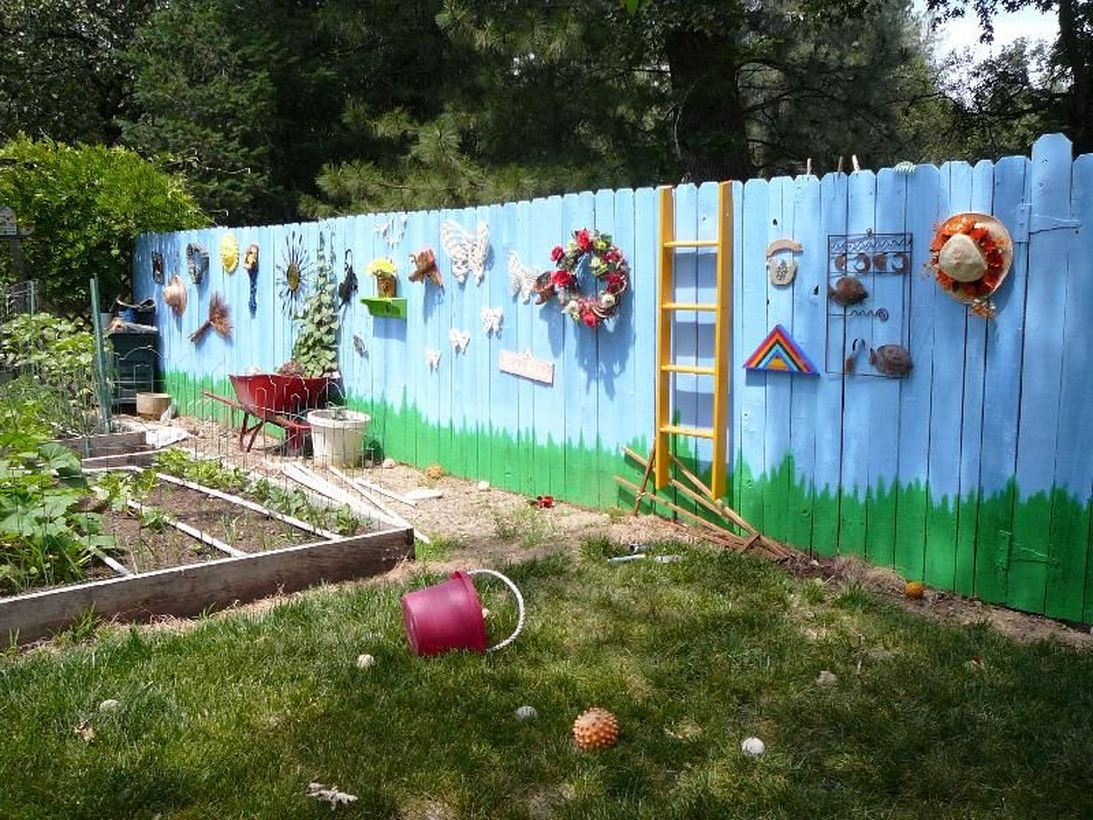 An adorable painting fence for garden with sky painted fence, grass and attach all kinds of knick-knacks hanging pleasant household appliances on the fence against a landscape background.