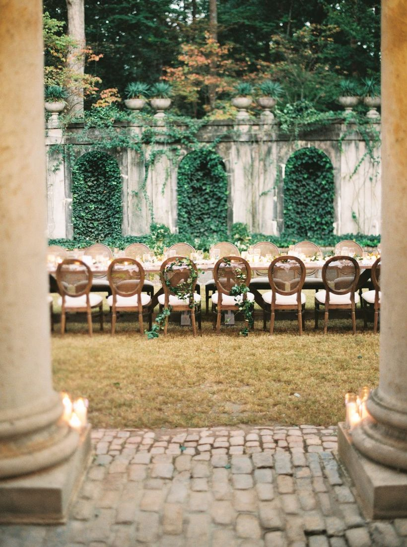 An amazing outdoor table set for wedding with a long wooden tables, wooden chairs, lush plants and crisp grass.