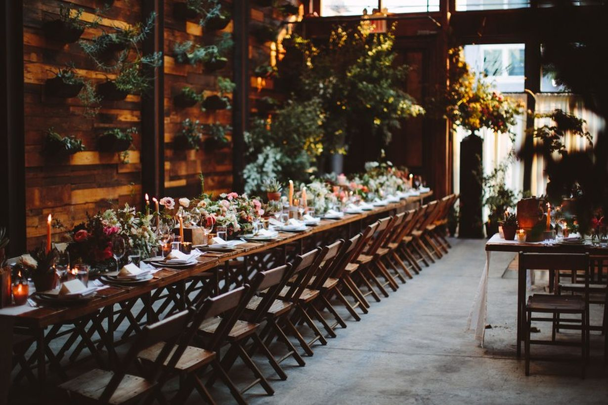 An attractive indoor table set for wedding with long wooden table decorated with colorful flower arrangements, mismatched candles, and eclectic dinnerware.
