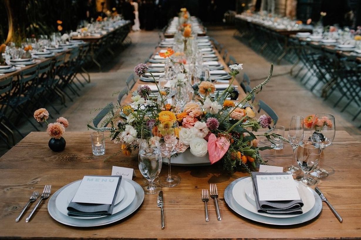 An incredible indoor table set for wedding with long bare wooden tables mirror the industrial and are dressed up modern tropical centerpieces in bright poppy colors.