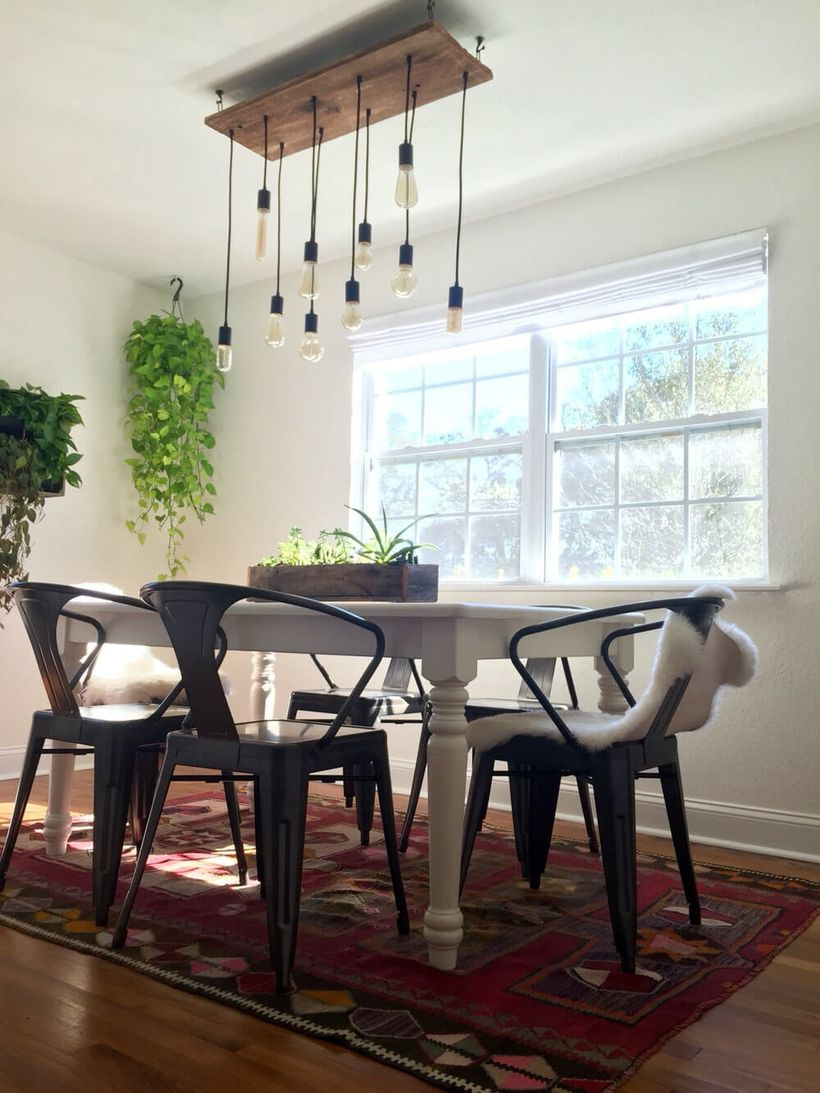 An exciting rustic chandelier for dining room with vintage bulb chandelier, a white table, iron black chairs, small carpet, house plants and big windows.