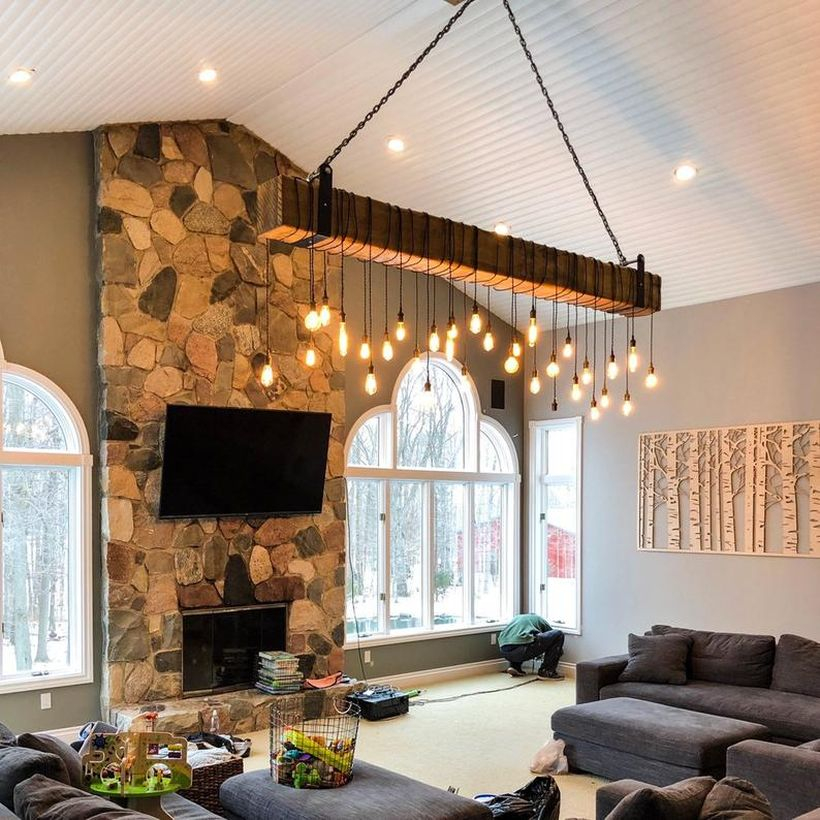 An interesting rustic chandelier for living room with long wooden beam chandelier, grey sofas, stone fireplace, large windows and decoration on the wall.