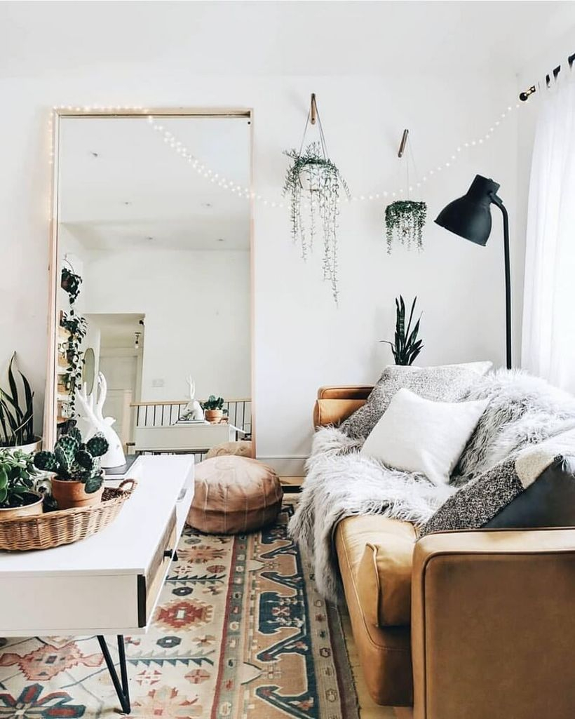 Boho living room design with white patterned carpet, long brown sofa, white and gray sofa cushion, square white coffe table, large mirror and hung green plants