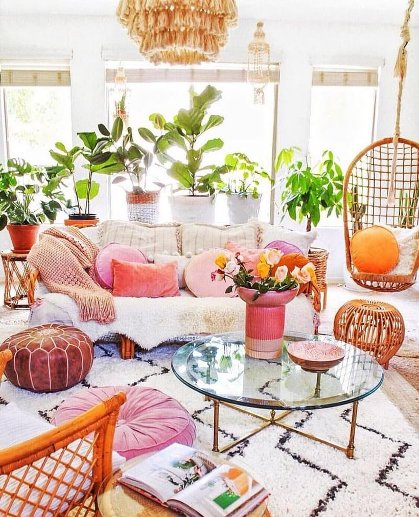 White boho living room design with white sofa, black stripe in white carpet, pink and white sofa cushions, hanging rattan chairs, round glass tables and plants in white fabric pots and rattan pots