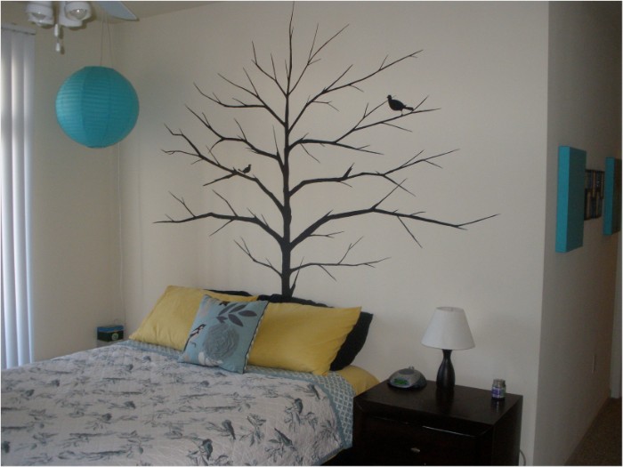 Mural-of-a-tree-with-bare-branches-and-two-birds-painted-in-black-on-a-cream-wall-near-a-bed-with-yellow-and-blue-cushions-and-a-dark-brown-bedside-table