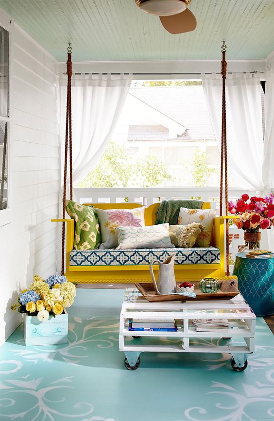 1-a-colroful-front-porch-with-a-yellow-hanging-bench-printed-pillows-a-pallet-coffee-table-and-some-blooms