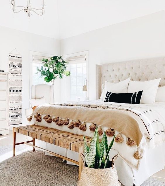 1-a-neutral-boho-bedroom-with-a-creamy-bed-a-woven-bench-boho-hangings-statement-plants-and-tassel-blankets