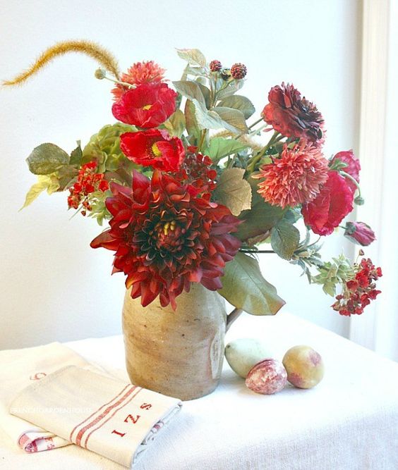 A-bright-fall-centerpiece-of-red-and-burgundy-blooms-greenery-in-a-vintage-jug-is-a-bold-decoration