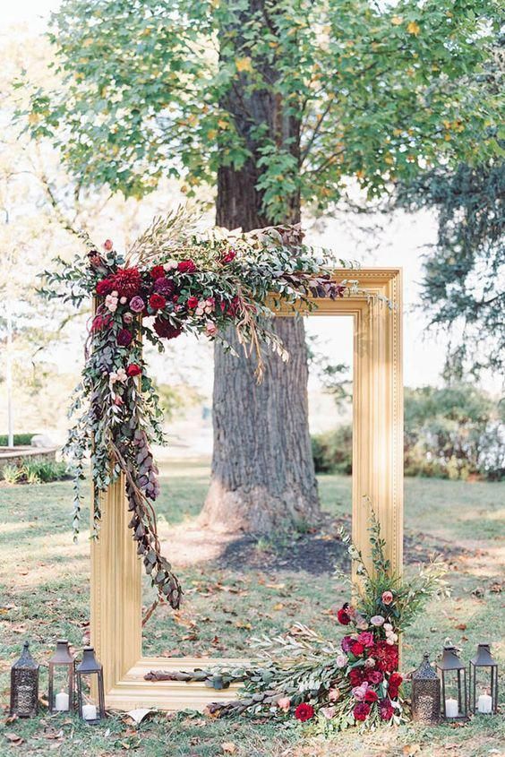 A-gold-frame-decorated-with-greenery-foliage-and-burgundy-blooms-plus-candle-lanterns-for-an-outdoor-fall-ceremony