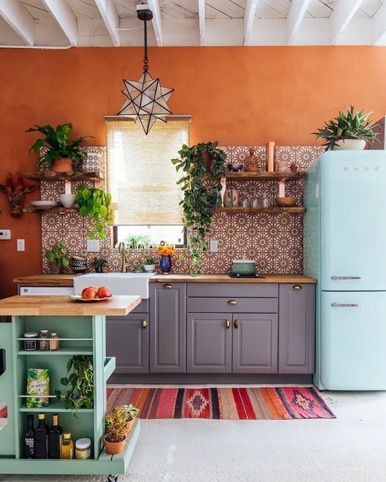 A-bright-kitchen-with-orange-walls-a-bright-printed-tile-backsplash-a-blue-fridge-a-green-kitchen-island-and-potted-greenery