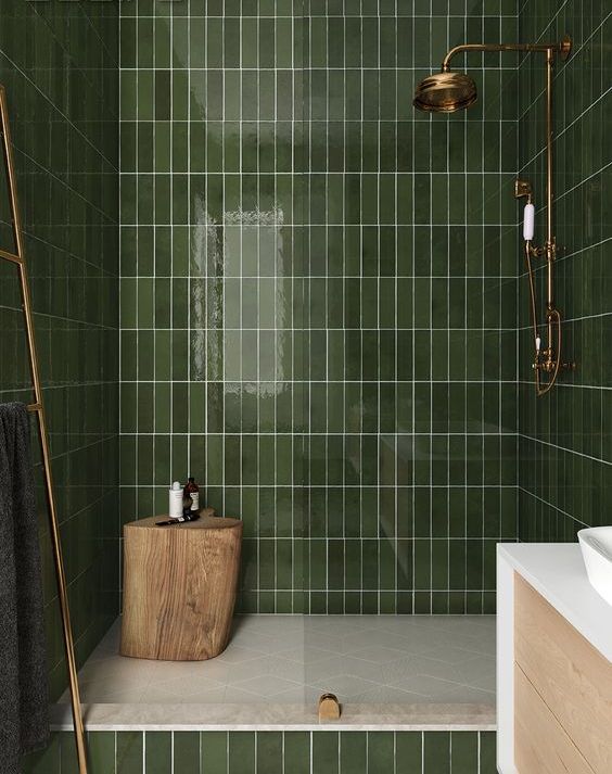 A-green-bathroom-clad-with-long-and-narrow-tiles-with-gold-and-brass-touches-and-a-tree-stump-stool-in-the-shower