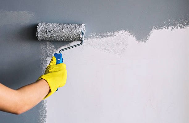Hand in yellow glove painting wall in gray color with a roller brush.