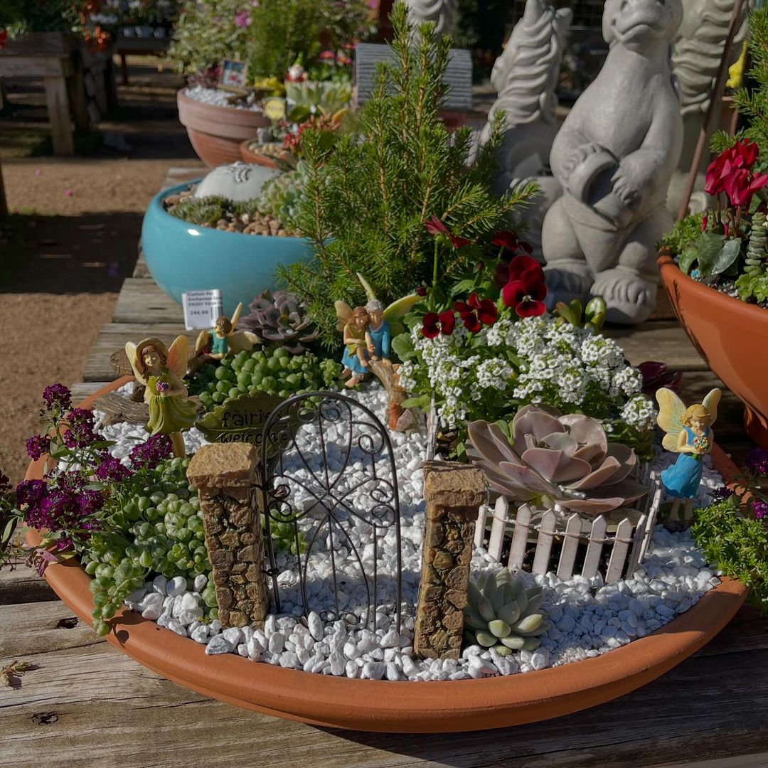 This fairy garden is set in a park where there is no home but just some plants, a seating area, plants, fence, and even a gate.